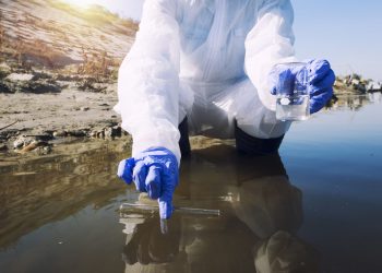 Unrecognizable ecologist taking samples of water with test tube from city river to determine level of contamination and pollution.